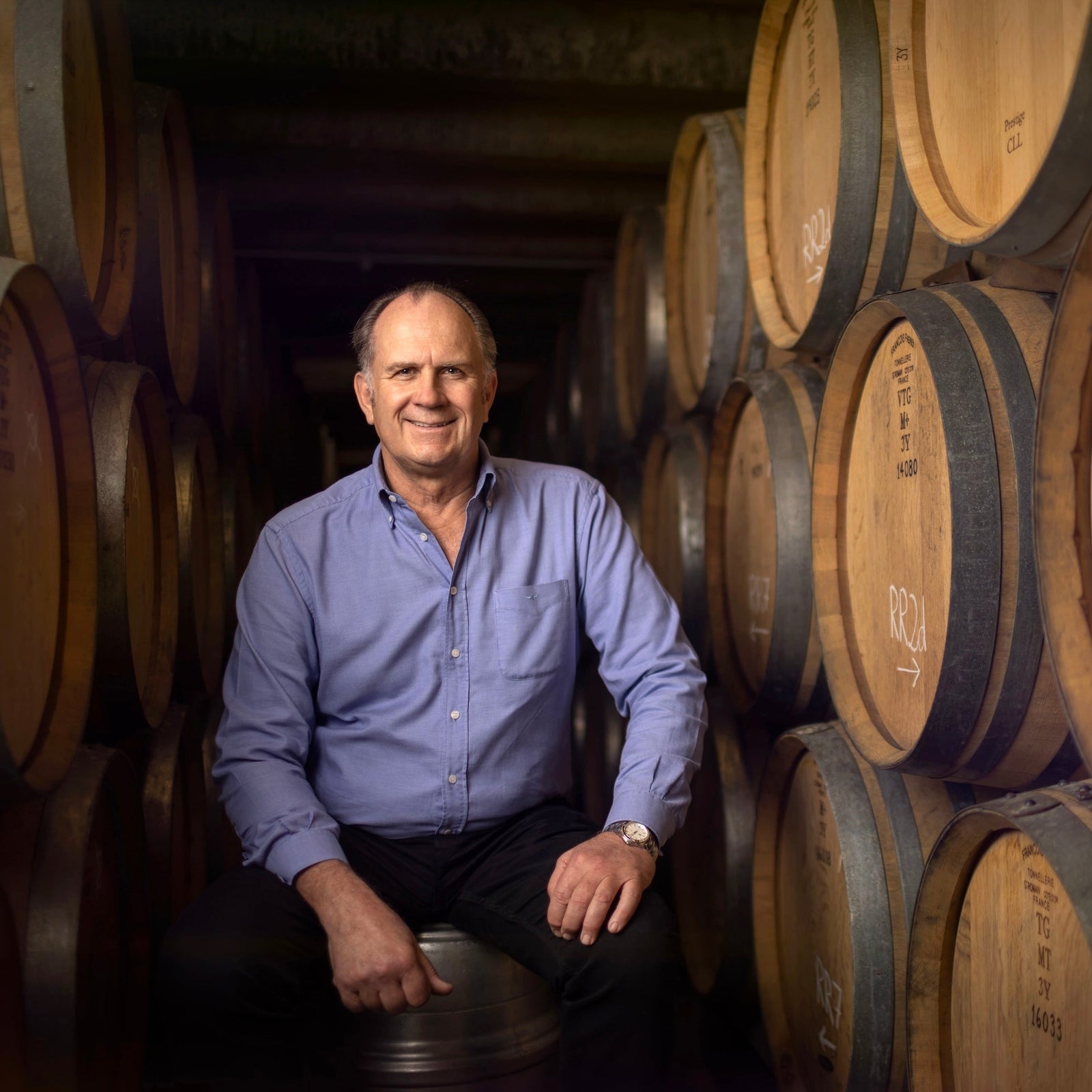 michael brajkovich in a light blue button-down shirt and dark trousers sits smiling between rows of large wooden wine barrels in a dimly lit cellar.